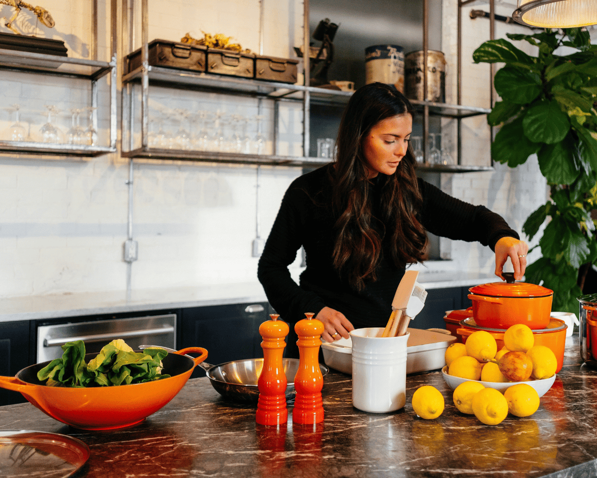 A woman in a black long-sleeve top stands in a modern kitchen preparing food, with orange cookware, lemons, and green salad on a dark marble countertop.